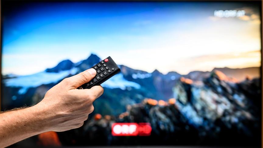 TV-Gerät, auf dem ORF-Progamm in HD zu sehen ist. Davor ist eine Hand zu sehen, die einen Knopf auf der Fernbedienung drückt.