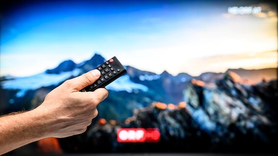 TV-Gerät, auf dem ORF-Progamm in HD zu sehen ist. Davor ist eine Hand zu sehen, die einen Knopf auf der Fernbedienung drückt.